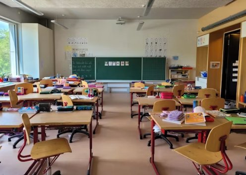 Aula vac&iacute;a con pupitres de madera en filas, una pizarra verde al fondo y material escolar encima de las mesas
