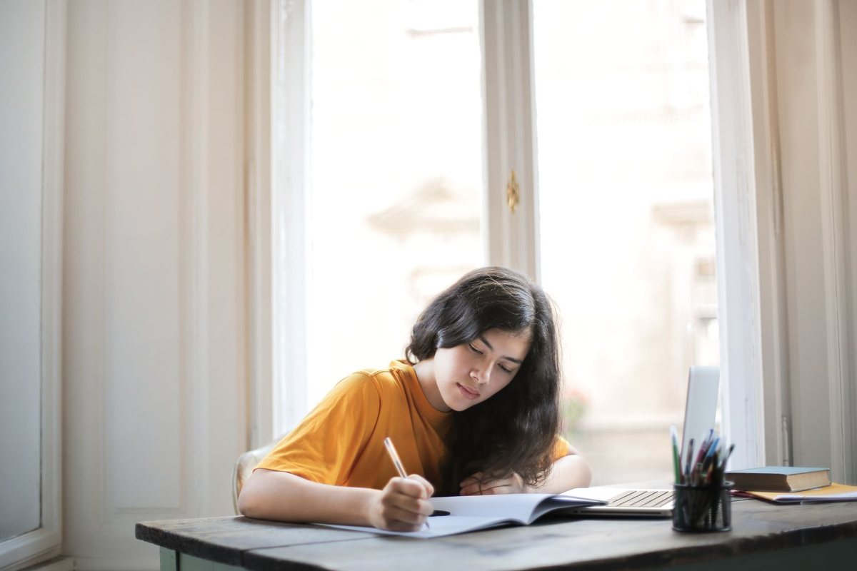 Una joven escribiendo a mano en un cuaderno junto a una ventana luminosa