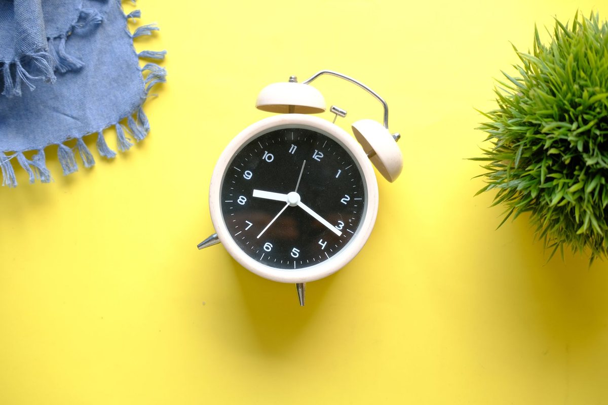 Reloj despertador crema sobre fondo amarillo, con una planta al lado